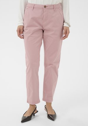Kobieta nosząca jasnoróżowe spodnie o prostych nogawkach z przednimi kieszeniami i zapięciem na guziki, w połączeniu z ciemnymi butami na obcasie z odkrytą piętą i zaostrzonym noskiem.