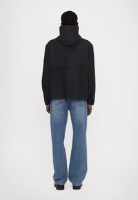 Jachetă neagră cu capotă, cu o croială lejeră și manșete elastice, combinată cu blugi evazați de culoare albastru deschis. Ambele articole prezintă un design minimalist.