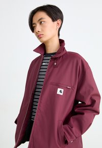 Bordeauxkleurige jas met een hoge kraag, voorzien van een rits aan de voorkant en een borstzak met een logo. Gestreept zwart-wit shirt eronder.