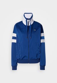 Jaqueta de fato de treino azul em poliéster com colarinho alto, fecho frontal, detalhes em branco e riscas. Apresenta um logótipo no lado esquerdo do peito.