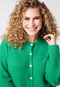 Groen gebreid vest met een ronde halslijn, voorzien van vijf witte knopen. De stof heeft een zachte, getextureerde uitstraling.