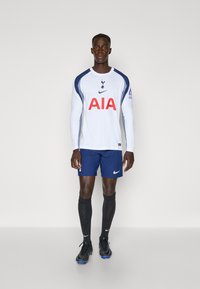 Hvid fodboldtrøje med lange ærmer, navy detaljer og rødt "AIA" logo, parret med navy shorts og sorte strømper, iført sorte støvler.