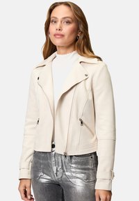 Giacca biker in suede finta crema con tasche con zip, rever a punta e silhouette aderente, abbinata a jeans argentati lucidi.