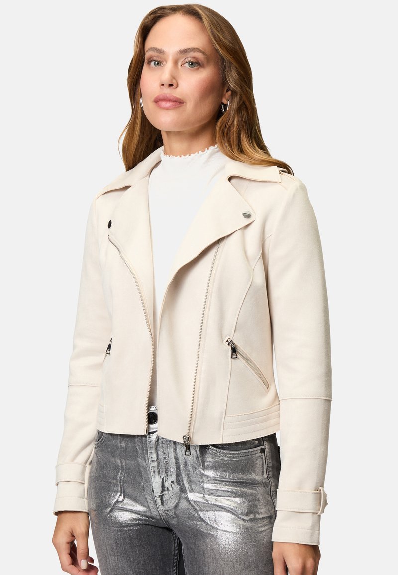 Giacca biker in suede finta crema con tasche con zip, rever a punta e silhouette aderente, abbinata a jeans argentati lucidi.