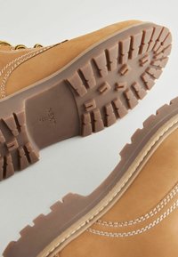 Botas de cuero color marrón claro con una suela de goma resistente que presenta un patrón de relieve texturizado. Costuras blancas acentúan el diseño lateral.
