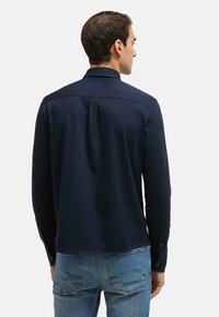 Dunkelblaues Langarmhemd aus glattem Stoff mit umklappbarem Kragen, einem Rückenteil und Knopfmanschetten, kombiniert mit hellblauen Jeans.