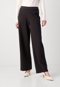 Vrouw in zwarte gestreepte wijde broek en beige mid-heel schoenen, van taille tot voeten tegen een effen achtergrond.