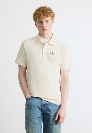 Noor punapea noormees, kes kannab kreemikat Calvin Kleini polosärki ja siniseid teksapükse, seisab tavalise valge tausta ees.