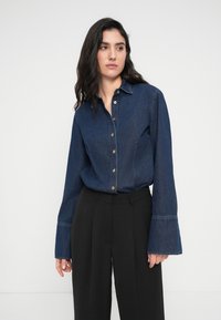 Σκούρο τζιν πουκάμισο με κουμπιά στο μπροστινό μέρος, μυτερό γιακά και φαρδιά μανίκια. Συνδυασμένο με ραμμένα μαύρα παντελόνια, δημιουργώντας μια αντίθετη εμφάνιση.