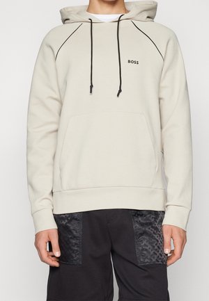 Person iført en beige hættetrøje med sorte snore og "BOSS" logo på brystet, kombineret med sorte shorts med mønstrede forlommer.