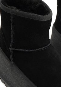 Melni potītes zābaki, kas izgatavoti no zamšādas ar mīkstu tekstūru, ar izliekta siluetu, melnu šuvumu un gumijas zoli.