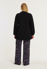 Kvinde med langt blond hår iført en sort langærmet top og brede sorte bukser med lilla og hvide blomster mønstre, stående med ryggen til.