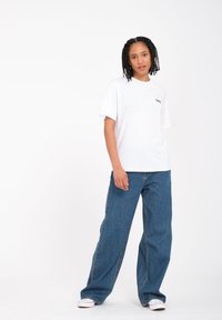 Weißes Baumwoll-T-Shirt mit kurzen Ärmeln und einem kleinen Logo auf der Brust, kombiniert mit lockeren blauen Jeans und weißen Sneakers.