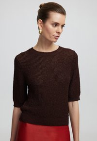 Suéter de punto marrón texturizado con mangas cortas y cuello redondo, combinado con una falda roja brillante. La tela tiene un patrón acanalado.