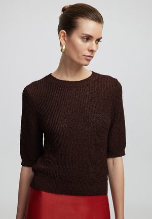 Maglione a maglia marrone con texture, maniche corte e scollo tondeggiante, abbinato a una gonna rossa lucida. Il tessuto presenta un motivo a coste.