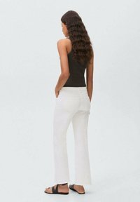 Black fitted tank top, white flared jeans with raw hem, and sandals with black accents. Model is viewed from behind, showcasing garment details.