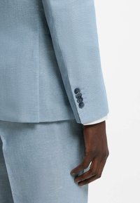 Main à la peau foncée à côté d'une manche de costume bleu clair avec quatre boutons et un pantalon assorti sur un fond blanc uni.