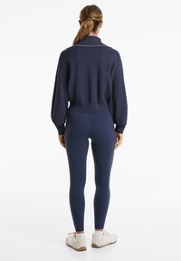 Dunkelblauer Strickpullover mit Kragen und weißem Streifen, kombiniert mit enganliegenden dunkelblauen Leggings und hellen Sneakers. Glatte Textur und einfaches Design.