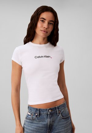 Kobieta z długimi ciemnymi włosami, ubrana w dopasowany biały t-shirt Calvin Klein i niebieskie dżinsy na jednolitym szarym tle.