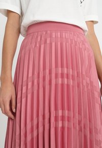 Mujer con una falda plisada rosa de talle alto con un sutil patrón a cuadros y una blusa blanca de manga corta.