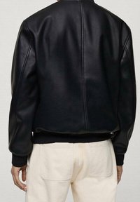 Personne portant une veste en cuir noire et un pantalon blanc cassé, vue de dos avec les mains détendues le long du corps.