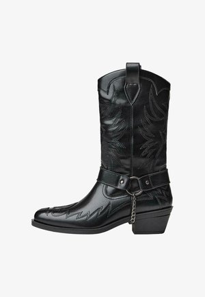 Stivale da cowboy in pelle nera con cuciture decorative, cintura, anello metallico, dettaglio in catena e tacco angolato basso su sfondo bianco.