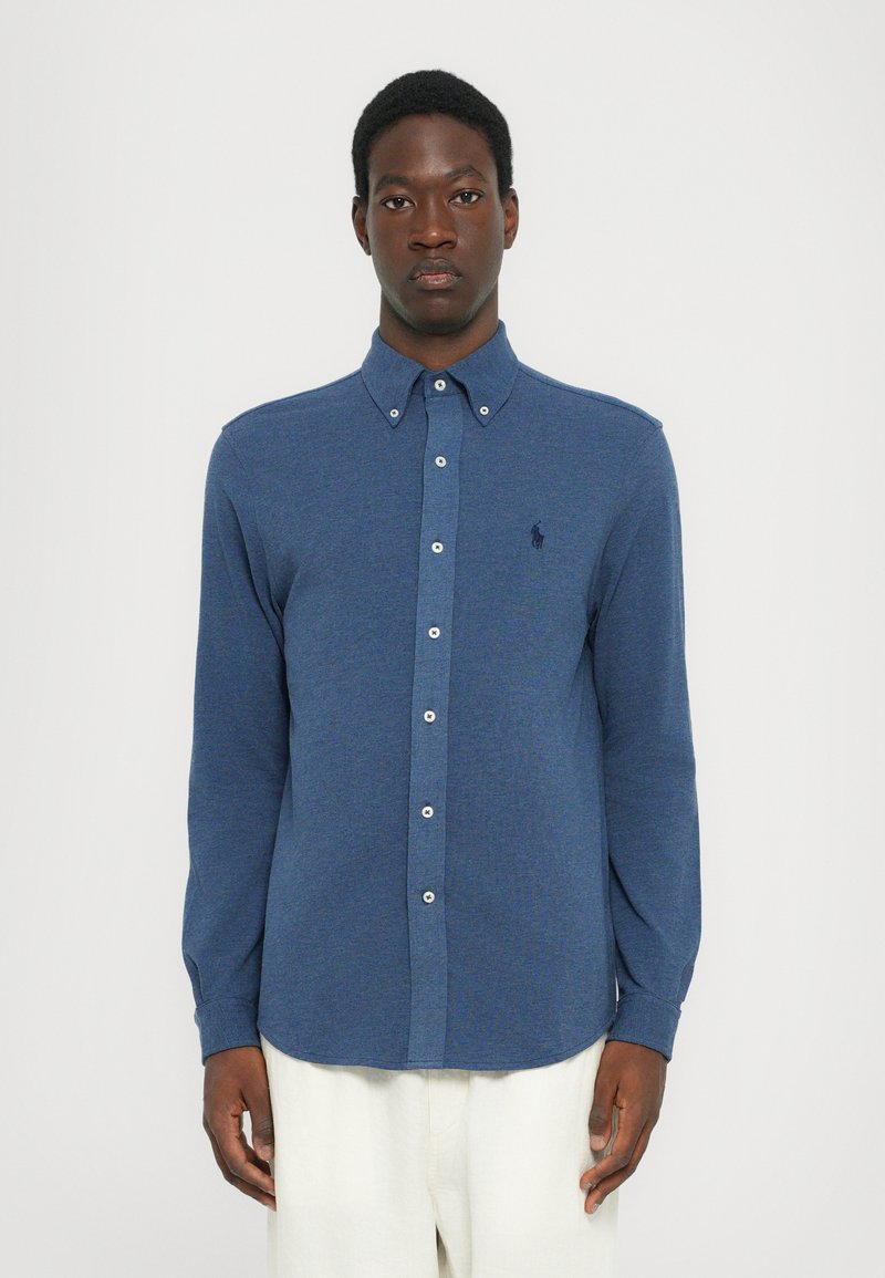 Chemise bleue à manches longues, boutonnée, en tissu texturé avec un col, dotée de boutons blancs et d'un petit emblème sur la poitrine.