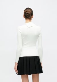 Λευκό πλεκτό πουλόβερ με μακριά μανί κια και γιακά, συνδυασμένο με μαύρη πλισέ φούστα. Το πουλόβερ διαθέτει υφασμένο ανάγλυφο ριμπ μοτίβο.