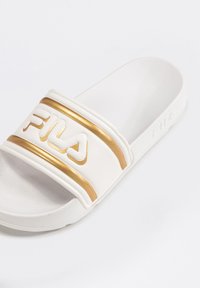 Chausson en slide blanc avec deux bandes dorées et logo "FILA" embossé sur la bride. Fabriqué en matériau lisse et texturé avec une semelle plate.