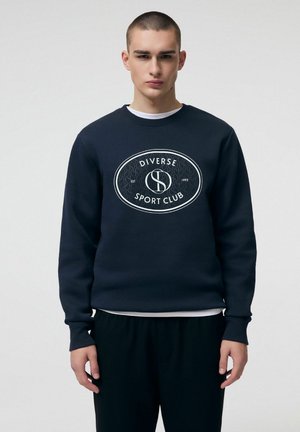 Młody mężczyzna z krótkimi włosami, ubrany w granatową bluzę z logo "Diverse Sport Club" i czarne spodnie, stojący na jednolitym tle.