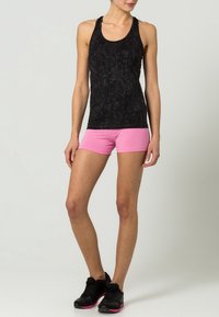 Czarny top na ramiączkach z teksturowanym wzorem w połączeniu z różowymi szortami. Modelka nosi czarne buty sportowe, stoi na neutralnym tle.