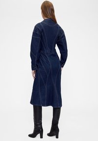 Tumesinises toonis teksakleit kraega, pikkade varrukate ja vöösse tõmmatud taljega. Omaduseks on kontrastne õmblus ja keskmise pikkusega disain.