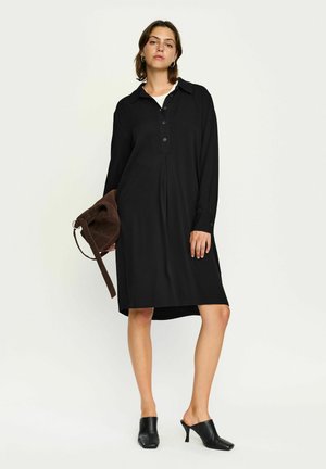 Vestito lungo nero a maniche lunghe con colletto a bottoni, patta frontale e design plissettato. Indossato con mule con tacco e borsa a spalla marrone.
