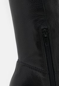 Botte montante en cuir noir avec une forme incurvée, une finition texturée et une fermeture éclair latérale avec une tirette noire. Coutures détaillées visibles.