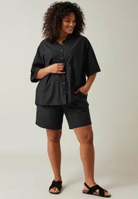 Camicia nera con bottoni e maniche corte, abbinata a pantaloni corti neri. Il modello indossa sandali neri con dettagli in sughero, su uno sfondo neutro.