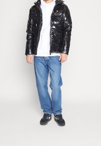 Černá lesklá prošívaná bunda s kapucí, předním zipem a dvěma kapsami, kombinovaná s bílou mikinou s kapucí, modrými džínami a černými teniskami.