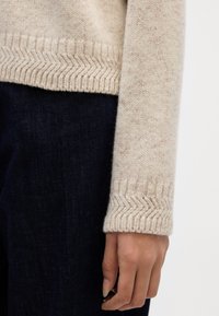 Beige gebreide sweater met een gewatteerde manchet in een visgraatpatroon. De stof lijkt zacht en warm, in combinatie met donkere denim jeans.