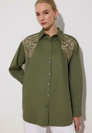 Žena v olivovo zelenej košeli na gombíky s béžovou kvetinovou výšivkou na ramenách a veľkými striebornými okrúhlymi náušnicami.