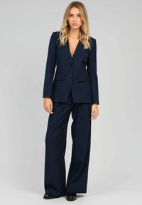 Femme portant un tailleur pantalon bleu marine avec des pantalons à jambes larges et un blazer boutonné, chaussée de chaussures en cuir noir, sur fond gris clair.