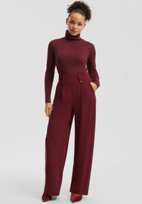 Bordeauxfarbener Rollkragenpullover kombiniert mit hochtaillierten, weit geschnittenen Hosen mit doppeltem Knopfdetail und seitlichen Taschen. Sanftes Stofffinish.