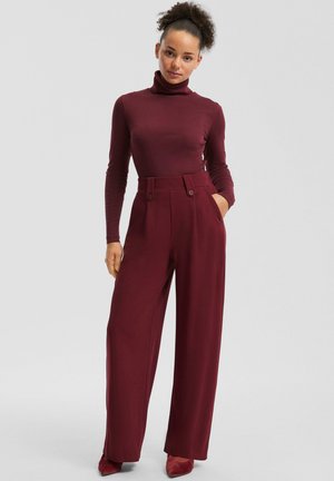 Bordeauxfarbener Rollkragenpullover kombiniert mit hochtaillierten, weit geschnittenen Hosen mit doppeltem Knopfdetail und seitlichen Taschen. Sanftes Stofffinish.