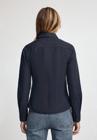 Camicia blu navy con maniche lunghe, collo a camicia e texture liscia. Presenta un taglio dritto e un design aderente sul retro.