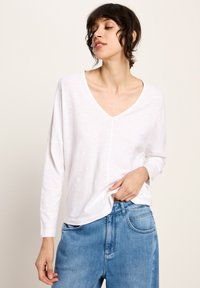 Frau mit kurzen dunklen Haaren, die ein lässiges weißes V-Ausschnitt-langärmliges Oberteil und blaue Jeans trägt, während sie den Saum des Shirts mit einer Hand hält.