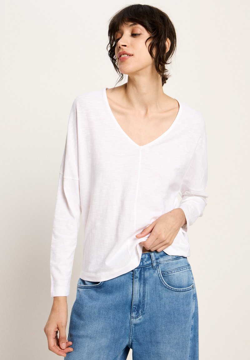 Frau mit kurzen dunklen Haaren, die ein lässiges weißes V-Ausschnitt-langärmliges Oberteil und blaue Jeans trägt, während sie den Saum des Shirts mit einer Hand hält.