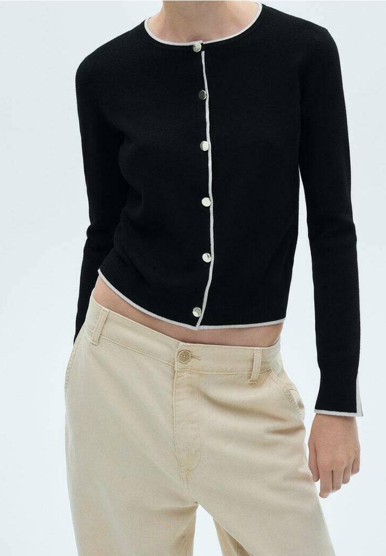 Personne portant un cardigan noir ajusté à manches longues avec bordure et boutons blancs, associé à un pantalon beige taille haute.