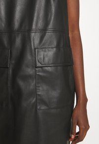 Vestito in pelle nera con una texture liscia, dotato di due tasche a toppa frontali e dettagli di cucitura ben visibili.