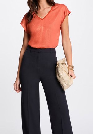 Orange kortærmet V-hals top med en subtil glans kombineret med højtaljede, vide sorte bukser; holder en guldfarvet clutch.