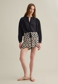 Schwarze Bluse mit kurzen Puffärmeln, kombiniert mit cremefarbenen und schwarzen, gemusterten Häkelshorts mit gewelltem Saum. Flache Sandalen runden das Outfit ab.