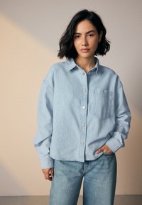 Light blue, oversized button-up shirt with a textured fabric, a chest pocket, and a classic collar, paired with light blue denim jeans.