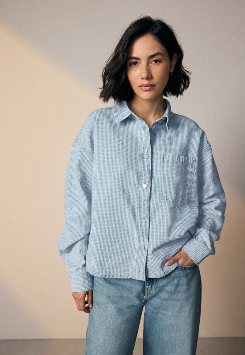 Light blue, oversized button-up shirt with a textured fabric, a chest pocket, and a classic collar, paired with light blue denim jeans.